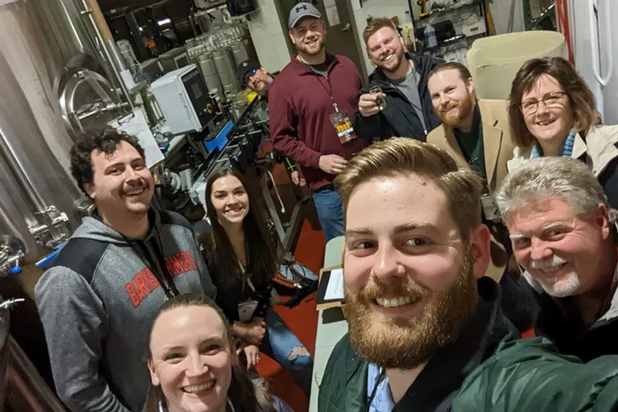 A group of smiling people is taking a selfie in a brewing facility surrounded by stainless steel equipment.