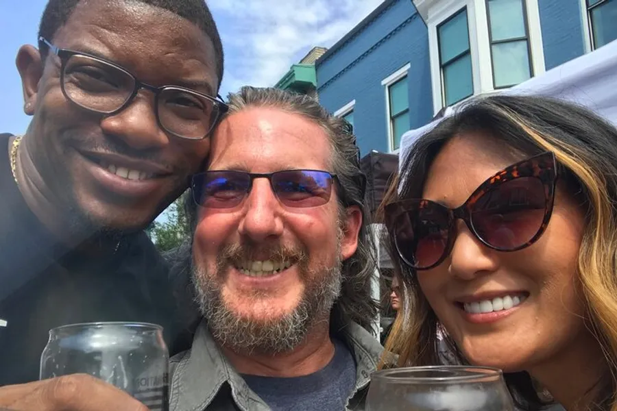 Three smiling adults are closely posing for a selfie at an outdoor event, holding drinks in their hands.