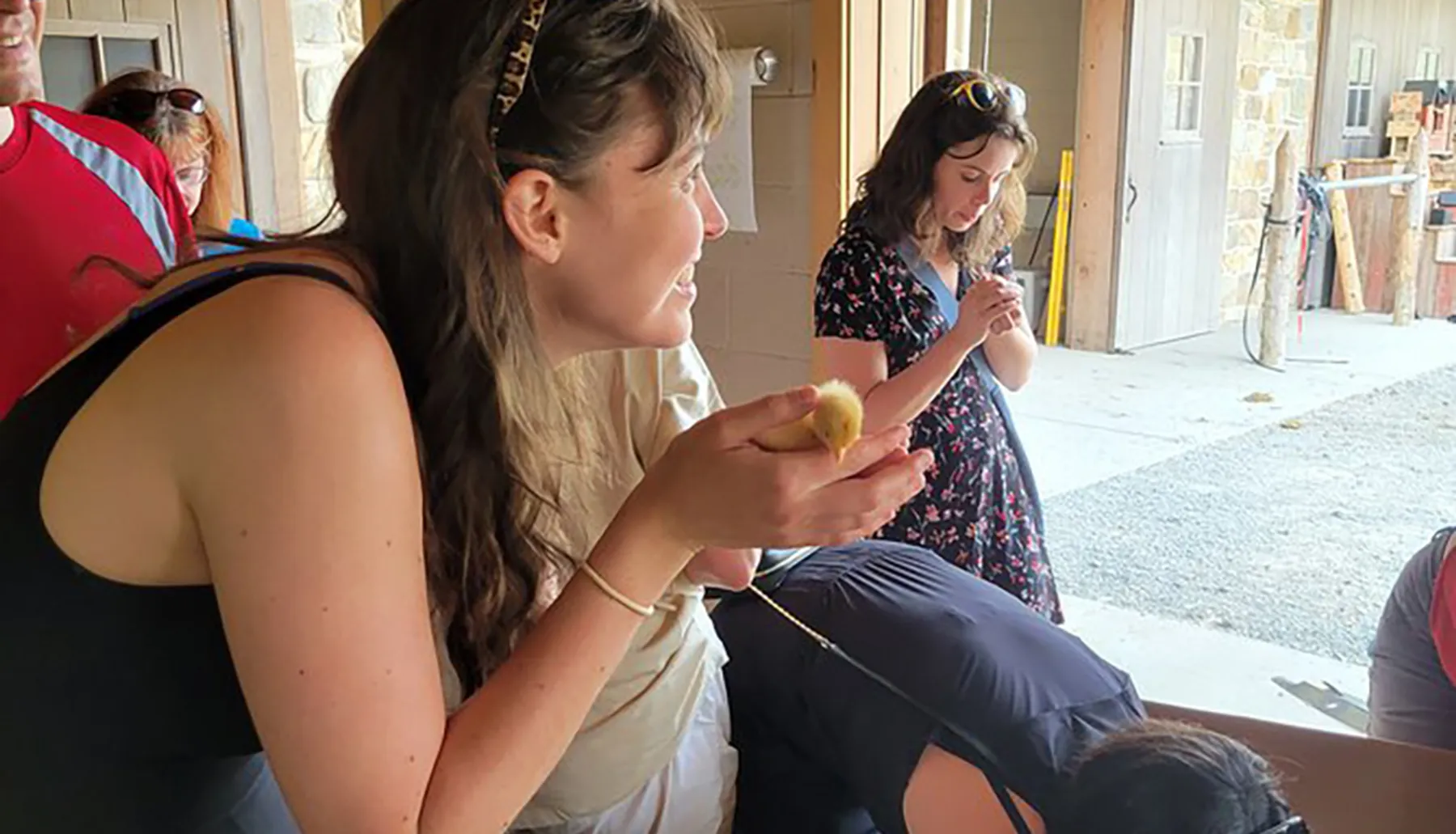 A woman is smiling and holding a small chick in her hands while another person is focused on her phone in the background, all in a rustic setting that appears to be a barn or farm.