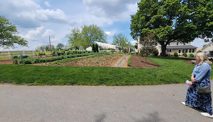A person is sitting on the curb of a paved path, looking at a lush vegetable garden next to a white residential building on a sunny day.