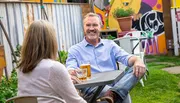 A man with a pleasant smile is enjoying a conversation and a glass of beer with a woman at an outdoor table in a colorful garden setting.
