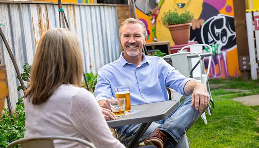 A man with a pleasant smile is enjoying a conversation and a glass of beer with a woman at an outdoor table in a colorful garden setting.