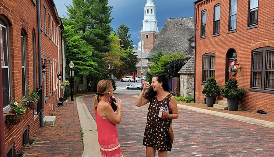 Two women are smiling and taking photos of each other on a quaint brick-lined street with historic buildings and a white church spire in the background.