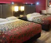 The image shows a hotel room with two double beds covered with multicolored patterned bedspreads bedside tables with lamps a chair a wall-mounted mirror and a framed picture creating a cozy sleeping area