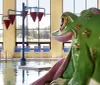 A large colorful frog-shaped sculpture is seated by an indoor pool with bright natural light streaming in from tall windows in the background