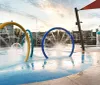 An empty water playground with colorful arches spraying water and a mushroom-shaped water feature with loungers and buildings in the background captured during the daytime