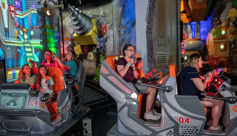 The image shows people on a ride, using blasters to interact with a colorful, game-like environment.