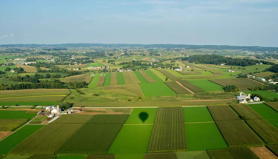 The image showcases a patchwork of lush green agricultural fields from an aerial perspective, with the shadow of a hot air balloon cast on the landscape.