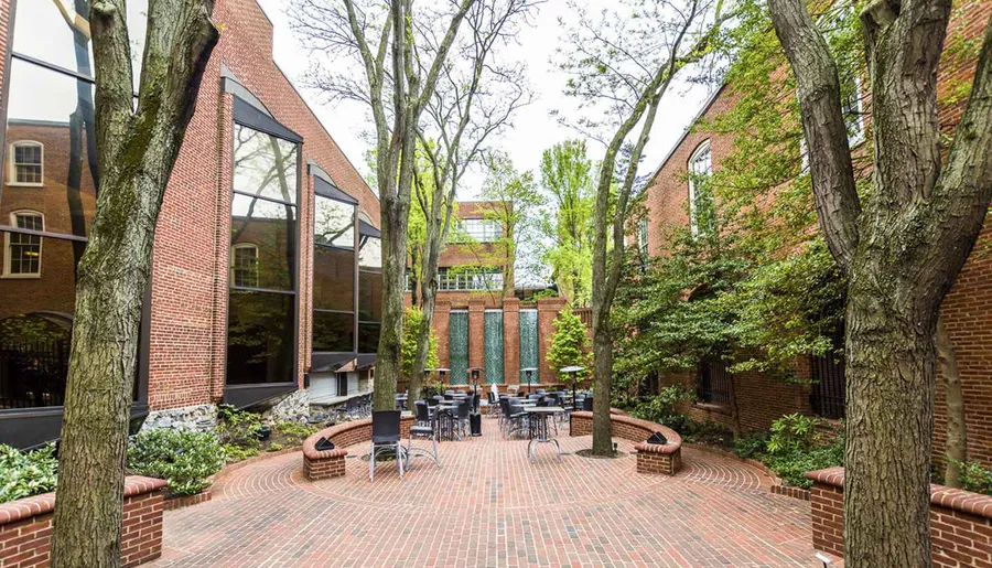 The image shows a serene outdoor patio area surrounded by brick buildings and tall trees, featuring tables and chairs for dining or relaxation.