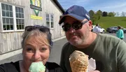 Two people are taking a selfie outside a creamery, each holding an ice cream cone and smiling on a sunny day.