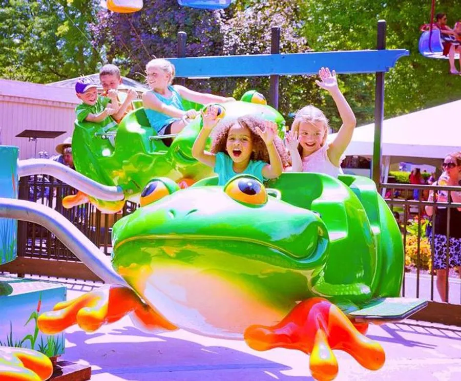 Children are joyfully riding on a colorful frog-themed amusement park ride on a sunny day.