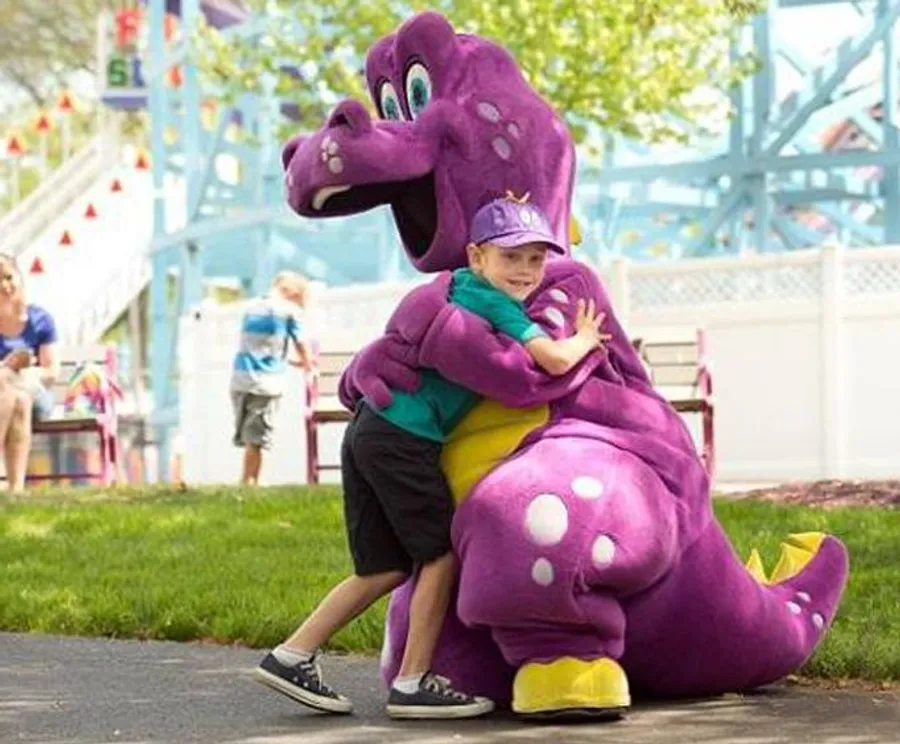 A child is joyfully hugging a person in a purple dinosaur costume at what appears to be an outdoor event or amusement park.