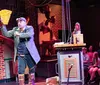 An individual in a steampunk-inspired costume holds up a large whimsical cheese wedge prop on stage entertaining an audience that includes both focused and distracted members