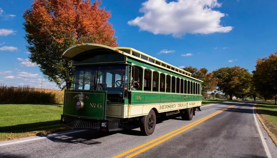 A vintage-style trolley bus labeled Hershey Trolley Works drives along a road flanked by trees with autumn foliage under a clear blue sky.