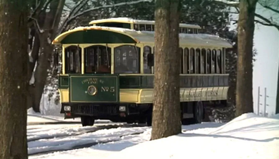 A vintage green and yellow tram is traveling through a snow-covered landscape flanked by trees.