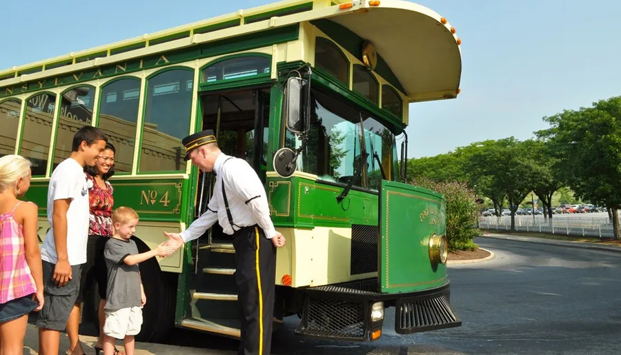 A trolley conductor is greeting a small child in front of a vintage-style green trolley with curious onlookers smiling nearby.