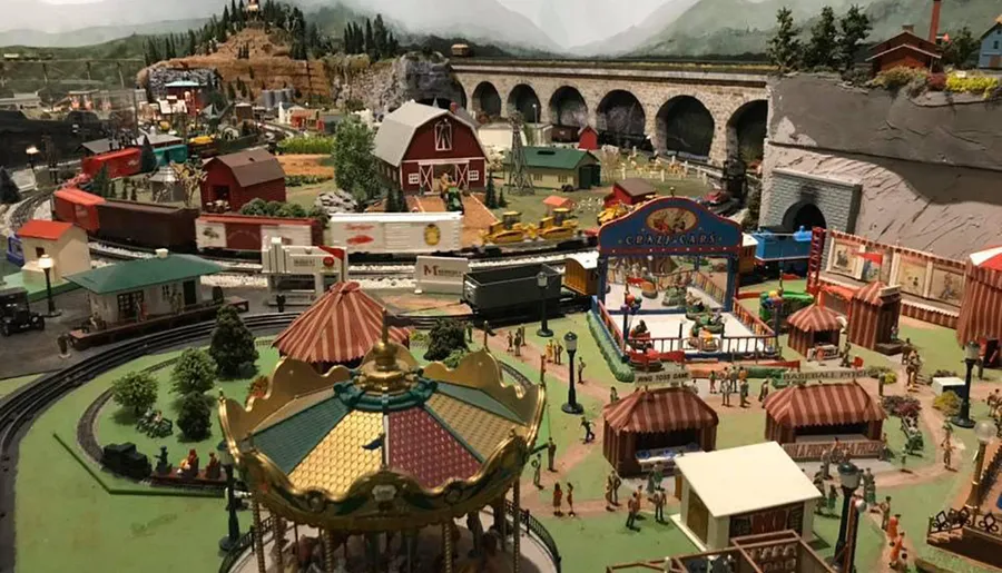The image shows a detailed miniature model layout featuring a vibrant carnival scene with a carousel, train tracks, various buildings, and small figurines representing people.