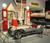 A classic convertible car is on display in a retro-themed exhibit with vintage gas pumps and signage in the background