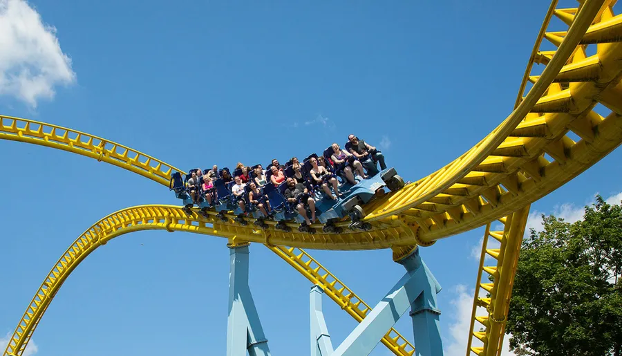 A roller coaster with excited riders is ascending a twisty yellow track against a clear blue sky with a few clouds.