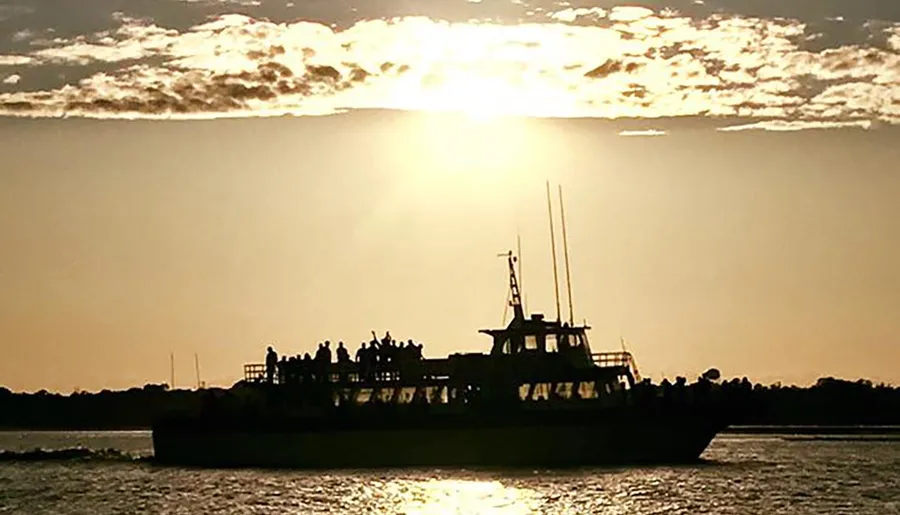 A ferry boat filled with passengers is silhouetted against a sunset backdrop over water.