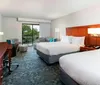 The image shows a tidy and modern hotel room with two queen-sized beds a desk area and a balcony