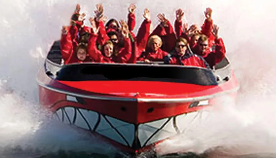 The image shows a group of excited passengers wearing red life jackets, raising their arms with joy while riding a high-speed boat creating large sprays of water.