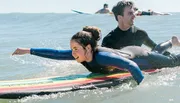 Two people are wearing wetsuits and lying on surfboards, paddling in the ocean.