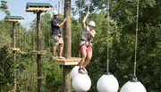 Two people are navigating an outdoor high ropes course among the trees, equipped with safety harnesses and helmets.