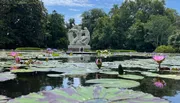 The image shows a tranquil pond with vibrant water lilies in the foreground and an imposing white sculpture of figures in the background, all set amidst lush greenery.
