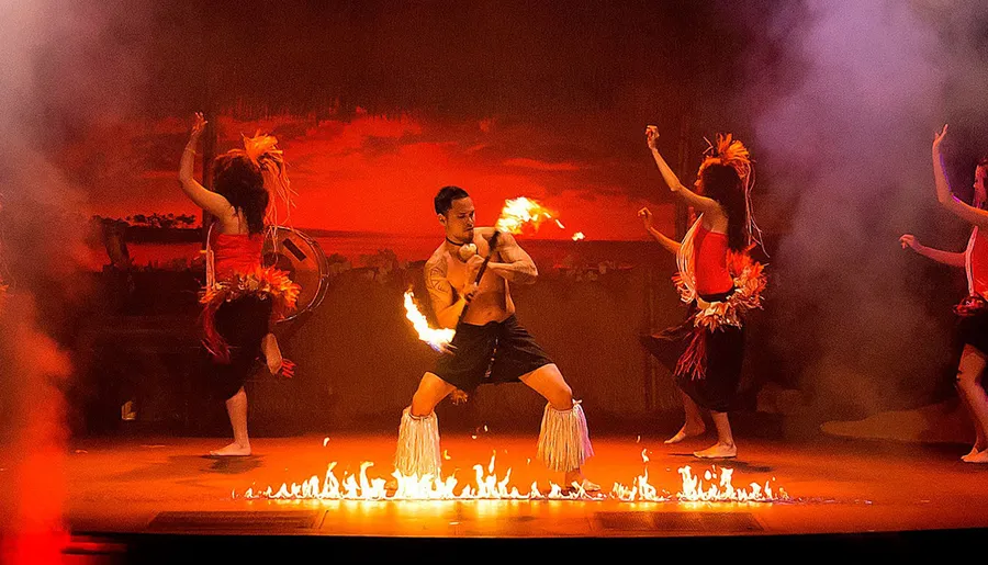 A group of performers engages in a traditional fire dance, with a central figure manipulating flames while others dance around him against a dramatically lit backdrop.