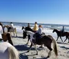 A group of people are horseback riding along a beach with a pier in the background