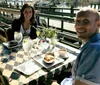 A man and a woman are smiling at the camera while enjoying a meal and drinks at an outdoor restaurant table with a water view in the background