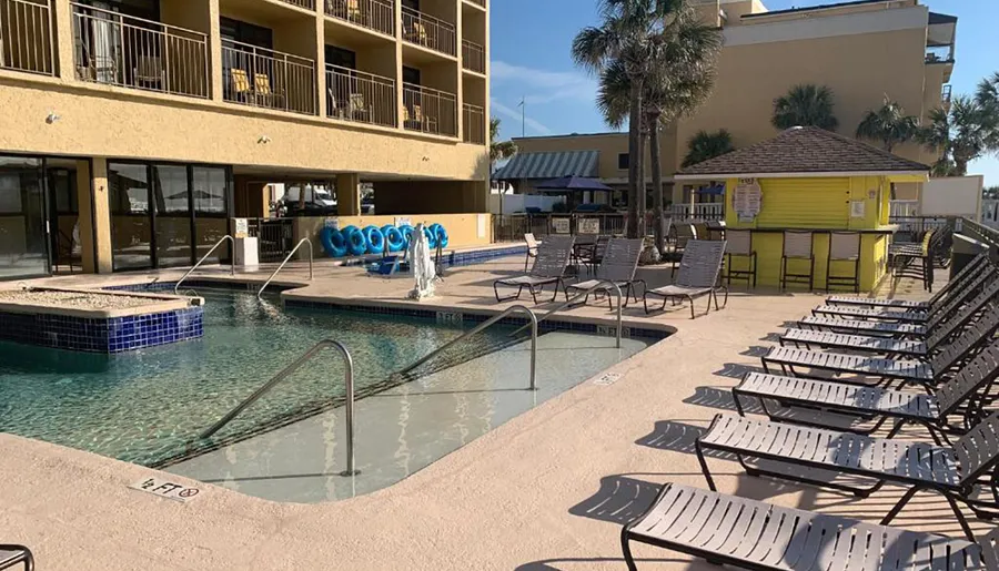 A hotel pool area with lounge chairs, blue float rings, and a small building, likely a pool bar or service area, under a clear sky.
