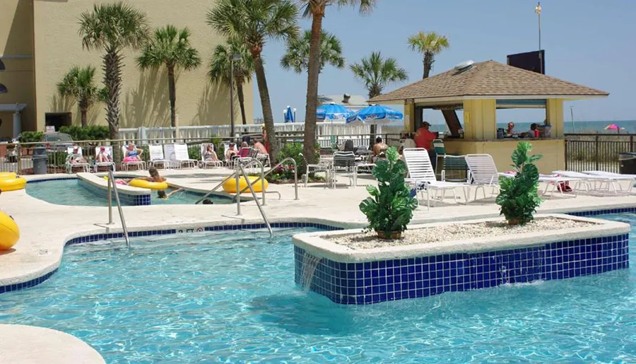 The image shows a sunny poolside area with people relaxing and swimming, deck chairs lined up, and a view of the beach in the background.