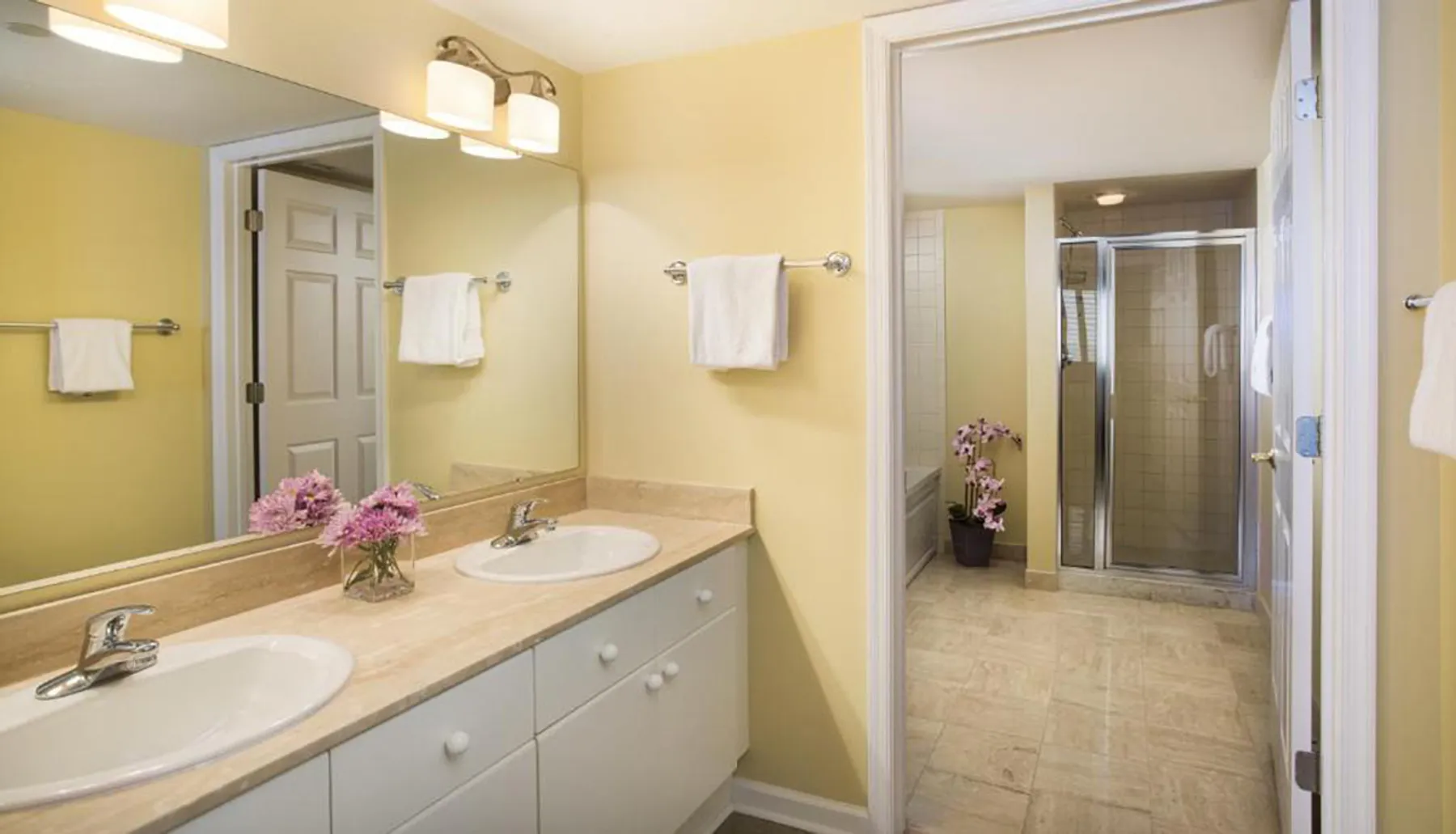 This image shows a tidy, yellow-painted bathroom with a double sink vanity, a large mirror with an overhead light fixture, and a walk-in shower visible through the open door.