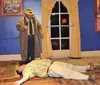 A man in a trench coat and hat appears to be investigating a scene where another person is lying motionless on the floor in what seems to be a staged play or a theatrical murder mystery setting