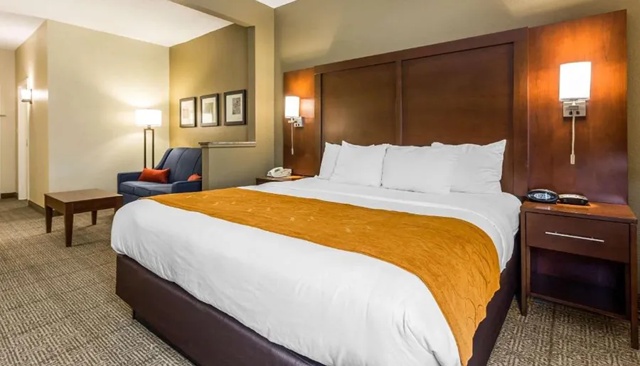 The image shows a neatly arranged hotel room with a large bed featuring white linens and an orange bed runner, accompanied by modern furnishings and decor.