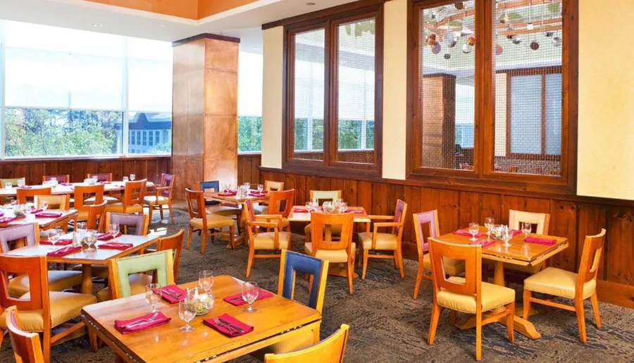 The image shows a well-lit, empty dining area with several wooden tables set for a meal, colorful napkins, and a view outside through large windows.