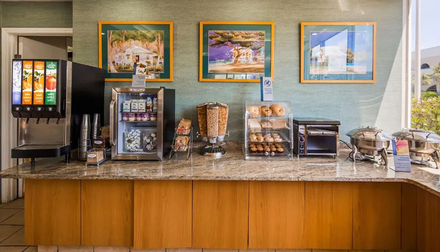 The image shows a hotel breakfast buffet with a selection of drinks and food including cereals, pastries, and hot items under chafing dishes, set against a wall adorned with colorful artwork.
