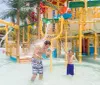 A child and an adult are enjoying playful moments in a splash water park with colorful water features and palm trees in the background