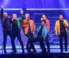 A group of six men are energetically performing on stage singing into microphones with dynamic expressions and colorful jackets