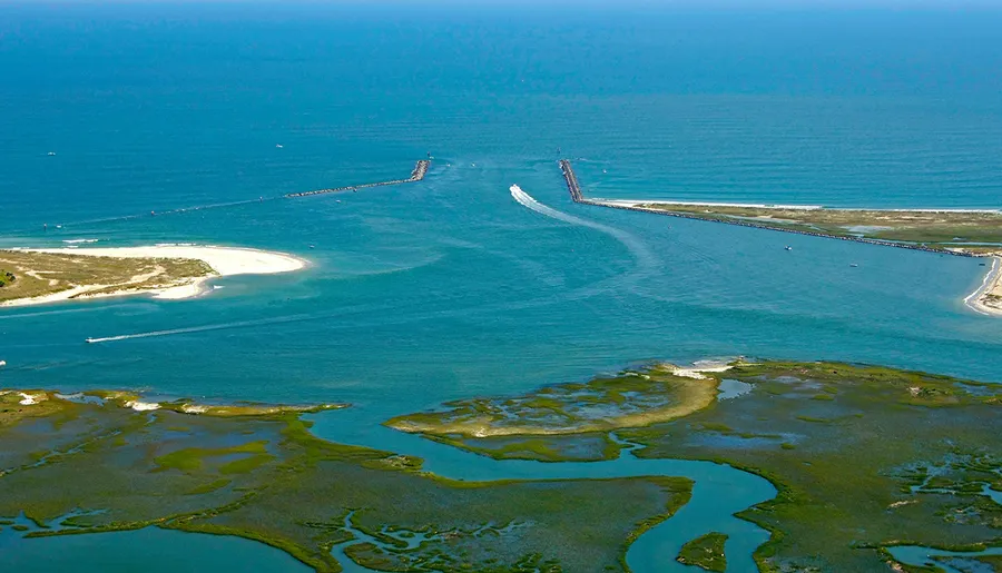 This is an aerial view of a coastal inlet with protective breakwaters, showing boats navigating the channel and surrounding wetlands.