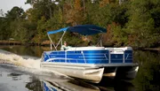 Two individuals are enjoying a ride on a blue and white pontoon boat with a blue sunshade on a calm river surrounded by trees.