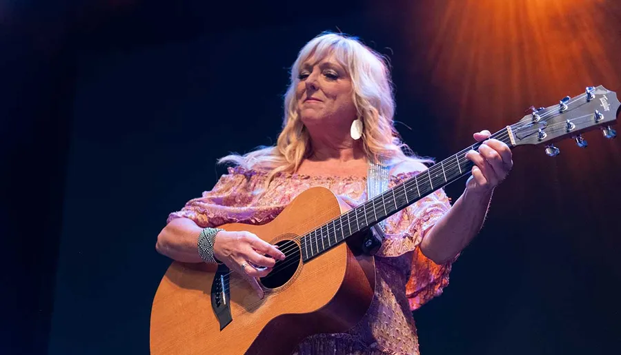 A woman is playing an acoustic guitar on stage under a warm spotlight.