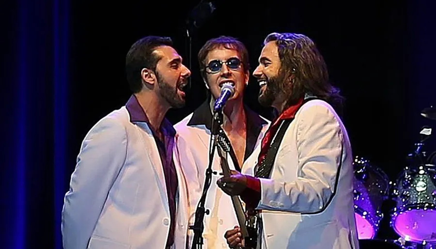 Three performers on stage are singing together into a microphone, dressed in white suits with a blue stage background and musical instruments behind them.