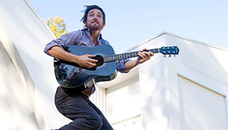 A person is energetically playing an acoustic guitar while appearing to be in mid-leap against a backdrop of a clear sky and a modern building.