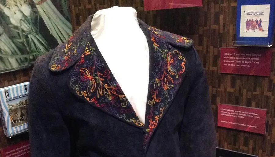 The image shows an elaborately embroidered jacket on a display mannequin with informational plaques in the background, suggesting it may be a piece of memorabilia from a musician or performer.