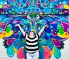 A person with arms raised stands in front of a vibrant colorful mural with abstract patterns and eye motifs