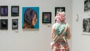 A person with pink hair and a whimsical outfit observes artwork displayed on a gallery wall.