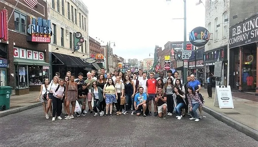 A diverse group of people poses for a photo on a street lined with music-themed stores and signs.